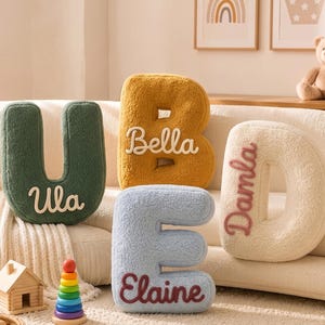 K&ouml;nnte beinhalten: Vier pl&uuml;schige buchstabenf&ouml;rmige Kissen in Dunkelgr&uuml;n, Senfgelb, Hellblau und Creme. Das gr&uuml;ne Kissen tr&auml;gt den Namen Ula, das gelbe Kissen Bella, das blaue Kissen Elaine und das cremefarbene Kissen zeigt den Namen Damla in Stickerei.