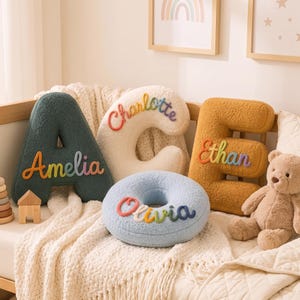 K&ouml;nnte beinhalten: Ein dunkelgr&uuml;nes Kissen in Form des Buchstabens A mit dem Namen Amelia, ein cremefarbenes C-Kissen mit Charlotte, ein senfgelbes E-Kissen mit Ethan und ein hellblaues rundes O-Kissen mit Olivia, alle aus strukturiertem Teddy-Stoff gefertigt.