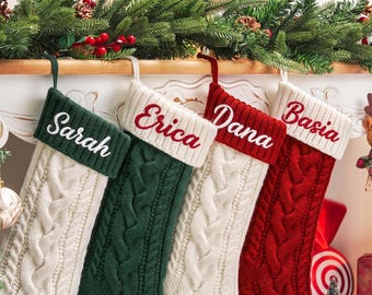 以下が含まれることがあります： ケーブルニットのクリスマスストッキングが4つ並んで吊るされています。1つ目は白で、ダークグリーンのカフにSarahという名前が刺繍されています。2つ目はダークグリーンで、白のカフにEricaと刺繍されています。3つ目は白で、赤のカフにDanaと刺繍されています。4つ目は赤で、白のカフにBasiaと刺繍されています。