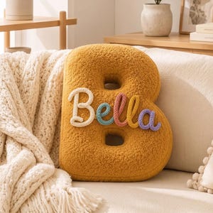 K&ouml;nnte beinhalten: Ein senfgelbes, strukturiertes Kissen in Form des Buchstabens B mit Sherpa-Optik. Der Name Bella ist mittig in Schreibschrift aufgestickt, wobei die einzelnen Buchstaben in den Farben Wei&szlig;, Blau, Rosa, Gelb und Violett gehalten sind.