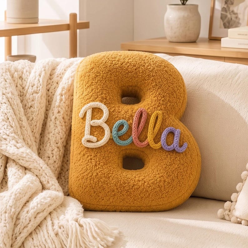 K&ouml;nnte beinhalten: Ein senfgelbes, strukturiertes Kissen in Form des Buchstabens B mit Sherpa-Optik. Der Name Bella ist mittig in Schreibschrift aufgestickt, wobei die einzelnen Buchstaben in den Farben Wei&szlig;, Blau, Rosa, Gelb und Violett gehalten sind.