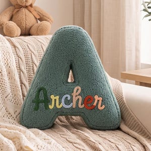 K&ouml;nnte beinhalten: Ein salbeigr&uuml;nes, A-f&ouml;rmiges Kissen aus strukturiertem Sherpa-Stoff. Auf der Vorderseite ist der Name Archer in bunten, einzelnen Buchstaben aufgestickt, wobei jeder Buchstabe einen anderen Farbton aufweist, darunter Dunkelgr&uuml;n, Gelb, Blau und Rot.