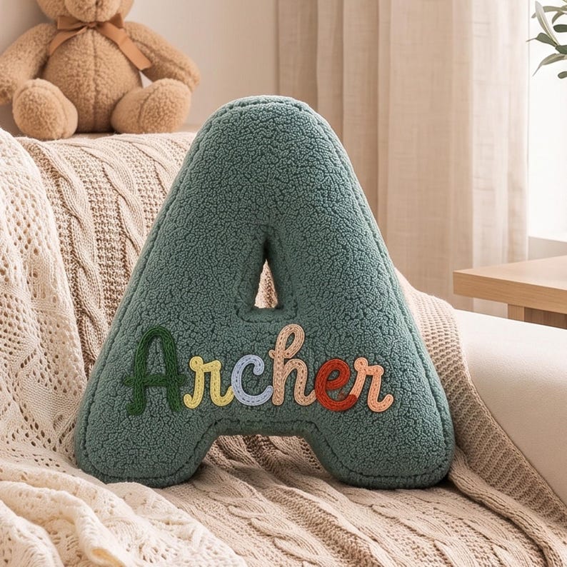 K&ouml;nnte beinhalten: Ein salbeigr&uuml;nes, A-f&ouml;rmiges Kissen aus strukturiertem Sherpa-Stoff. Auf der Vorderseite ist der Name Archer in bunten, einzelnen Buchstaben aufgestickt, wobei jeder Buchstabe einen anderen Farbton aufweist, darunter Dunkelgr&uuml;n, Gelb, Blau und Rot.