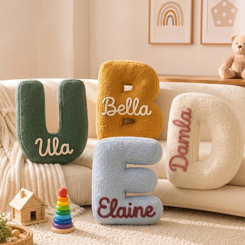 K&ouml;nnte beinhalten: Vier pl&uuml;schige buchstabenf&ouml;rmige Kissen in Dunkelgr&uuml;n, Senfgelb, Hellblau und Creme. Das gr&uuml;ne Kissen tr&auml;gt den Namen Ula, das gelbe Kissen Bella, das blaue Kissen Elaine und das cremefarbene Kissen zeigt den Namen Damla in Stickerei.