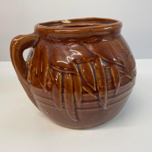 May include: A round, brown glazed ceramic pot featuring a single handle on the left side and a relief pattern of vertical leaves around the exterior. The rim has a small chip, and the surface has a glossy finish with horizontal ridges at the base.