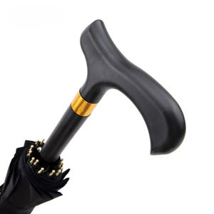 Pode incluir: Um guarda-chuva preto com um cabo curvo em forma de T e um anel de metal dourado localizado logo abaixo do cabo. O guarda-chuva é mostrado na posição fechada com o tecido reunido em torno do eixo central e pontas de cor dourada.