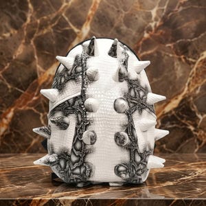 Puede incluir: Una mochila blanca y negra con un patrón de piel de serpiente texturizado y una serie de prominentes púas blancas puntiagudas dispuestas verticalmente a través del centro y los lados de la superficie exterior, creando un aspecto muy texturizado.