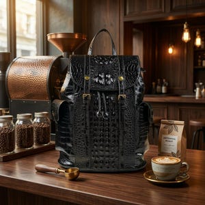 May include: A black backpack with a textured crocodile skin pattern, featuring a top handle, two vertical straps with gold-toned buckles, a drawstring closure, and two side pockets. The bag is positioned on a wooden surface next to a coffee cup.