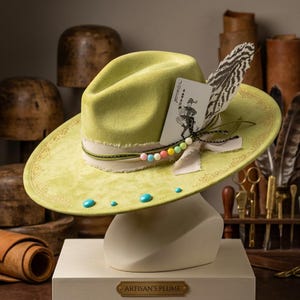 May include: A lime green felt hat featuring a wide brim with gold filigree detailing, a cream-coloured frayed ribbon band, a Joker playing card, a striped feather, a string of multicoloured beads, and several small turquoise-coloured cabochons.