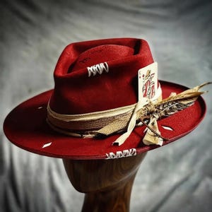 May include: A deep red fedora hat featuring a wide brim with white stitched detailing. The crown is adorned with a beige textured band, a King of Diamonds playing card tucked into the side, and a cluster of brown and white patterned feathers.