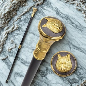 Peut inclure: Une canne de marche avec un fût noir et une poignée dorée surmontée d'une incrustation circulaire sombre présentant un relief de tête de loup en or. Un médaillon circulaire assorti avec le même motif de tête de loup est placé à côté de la canne.