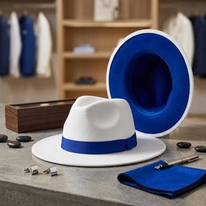 May include: A white fedora hat with a blue ribbon band, accompanied by a second fedora showing a blue interior, a blue pocket square, a silver fountain pen, and a pair of silver cufflinks. The items are arranged on a flat surface.