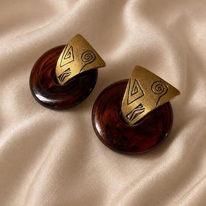 May include: A pair of earrings featuring circular, dark brown, marbled resin discs. Each disc is accented with a triangular, brushed gold-tone metal piece that features etched geometric lines and a spiral design on its surface.
