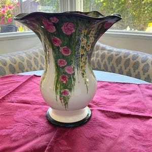May include: A ceramic vase with a flared, wavy rim and a glossy white finish. The exterior is hand-painted with vertical clusters of pink roses and trailing yellow and blue wildflowers against green foliage. The base and rim feature a black border.