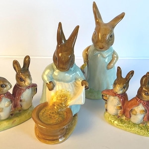 May include: A collection of ceramic rabbit figurines featuring two groups of three small bunnies in red capes on green bases, one standing rabbit in a blue coat, and one rabbit in a blue coat pouring yellow contents into a brown bowl.