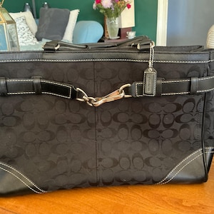 May include: A black Coach tote bag featuring the signature C monogram pattern on fabric. The bag includes black leather trim, white contrast stitching, a horizontal leather strap across the front with a silver-tone metal clasp, and a leather hangtag.