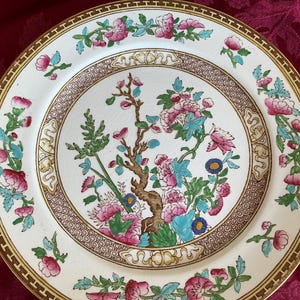 May include: A round ceramic plate featuring a central illustration of a brown tree with pink and blue flowers. The rim is decorated with a gold geometric border and a repeating pattern of pink flowers and green leaves on a cream background.