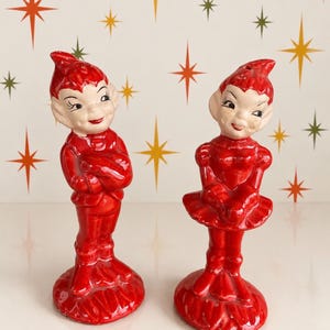 May include: Two glossy red ceramic elf figurines standing on circular bases. The left elf wears a jacket and pants with arms crossed. The right elf wears a dress with a flared skirt and puffed sleeves. Both have pointed ears and hats.