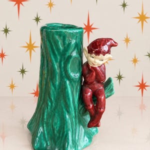 May include: A ceramic vase shaped like a green tree trunk with a glossy finish. A small, red-clad elf figure with a pointed hat and pale skin is perched on a branch extending from the side of the trunk, resting its chin on its hands.