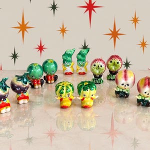 May include: A collection of twelve small ceramic salt and pepper shakers shaped like anthropomorphic fruits and vegetables. Each piece features a hand-painted face, colourful glossy glaze, and small holes on top for dispensing seasoning.