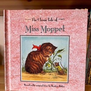 May include: A hardcover book titled The Classic Tale of Miss Moppet, based on the original story by Beatrix Potter. The cover features a pink textured background and a central illustration of a brown tabby kitten with a red bow and a mouse.