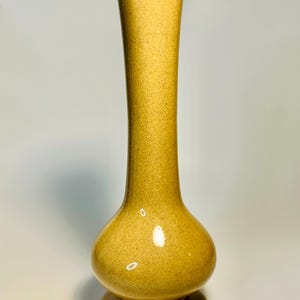 May include: A tall, slender ceramic vase with a mustard yellow speckled glaze. The vase features a bulbous, rounded base that tapers into a long, narrow neck, ending in a simple circular opening. The surface has a smooth, glossy finish.