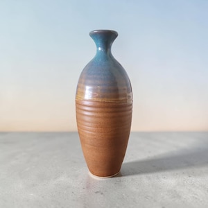 Peut inclure: Un vase en céramique avec un col étroit et un corps arrondi. La partie supérieure présente un émail bleu lisse et brillant, tandis que la section inférieure est finie en argile brune mate et texturée avec des stries horizontales.