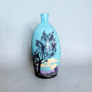May include: A light blue ceramic vase with a narrow neck and rounded body. The surface features a painted landscape scene with a large black silhouette of a tree in the foreground, soft yellow clouds, and a purple horizon line against a blue sky.