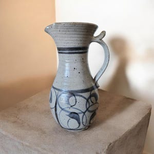 May include: A ceramic pitcher with a light grey speckled finish, featuring a dark grey horizontal band around the neck and dark grey swirling patterns around the base. The pitcher has a curved handle and a small spout at the top rim.