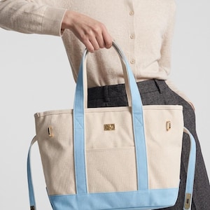May include: A beige canvas tote bag featuring light blue trim, two sturdy top handles, and a rectangular gold-toned metal plaque centered on the front. The bag includes side straps with gold-toned hardware and an open exterior pocket.