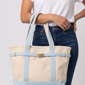 May include: A cream-colored canvas tote bag featuring light blue trim, dual shoulder straps, and a horizontal light blue strap across the front. The bag includes a gold-toned metal turn-lock closure centered on the front strap and a front pocket.