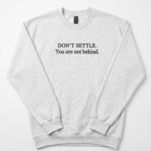 May include: A light gray heathered crewneck sweatshirt featuring the black printed text DON'T SETTLE. You are not behind. centered on the chest. The garment has long sleeves with ribbed cuffs and a ribbed neckline, providing a classic pullover style.