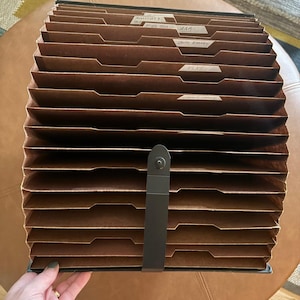 May include: A brown accordion-style expanding file folder with multiple dividers and tabs. The tabs feature handwritten labels including AAA, Smith Barney, Elks, and Credit Union. A black vertical strap with a snap closure secures the folder.