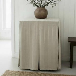 May include: A square table covered by a floor-length, light beige tablecloth featuring vertical white pinstripes. A dark brown, rounded ceramic vase sits on top of the table, containing a cluster of thin, bare, dark-colored decorative branches.