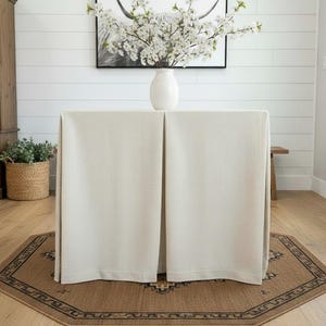 May include: A rectangular table covered with a solid cream-colored floor-length tablecloth. A white ceramic vase filled with white cherry blossom branches sits on top. The table is placed on an octagonal brown rug with a black geometric border.