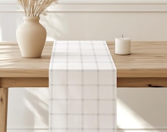 May include: A white table runner with a subtle gray plaid pattern. The runner is laid on a wooden table with a vase of dried grass and a white candle.