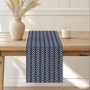 May include: A navy blue table runner with a white chevron pattern. The runner is on a wooden table with a vase of dried grass.