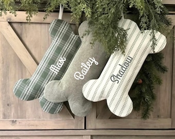 May include: Three personalized dog bone stockings with names Max, Bailey, and Shadow. The stockings are made of a plaid fabric, a gray fabric with white stripes, and a white fabric with green stripes.