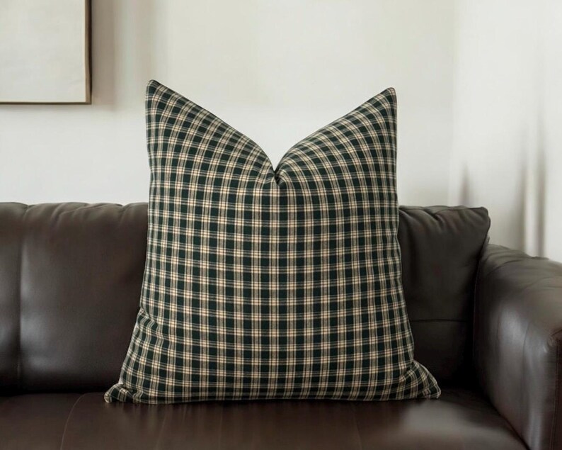 May include: A green and cream plaid throw pillow with a square pattern. The pillow is sitting on a brown leather couch.
