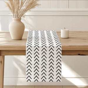 May include: A white table runner with a black chevron pattern. The runner is on a wooden table with a white vase and a candle.