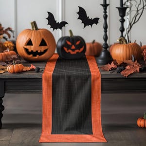 May include: A Halloween-themed table setting featuring a black and orange table runner. The scene includes carved pumpkins, decorative bats, and fall foliage. The pumpkins are orange and black, with carved jack-o'-lantern faces.