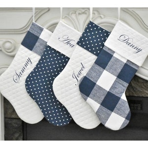 May include: Four white Christmas stockings with navy blue accents. Each stocking has a different pattern: plaid, polka dots, and quilted. Names Sammy, Ava, Jewel, and Danny are embroidered on the stockings.