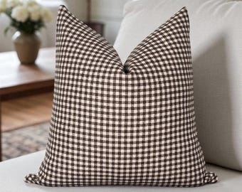 May include: A decorative throw pillow with a brown and white gingham pattern. The pillow is square with a pointed top and is resting on a white sofa. The pillow adds a touch of rustic charm to any living space.