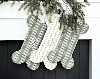 May include: Three dog bone-shaped Christmas stockings in green and white patterns hang from a white mantel. The stockings are personalized with the names "Sully", "Rudi", and "Yogi" in white script. Green pine branches and pine cones are above the stockings.