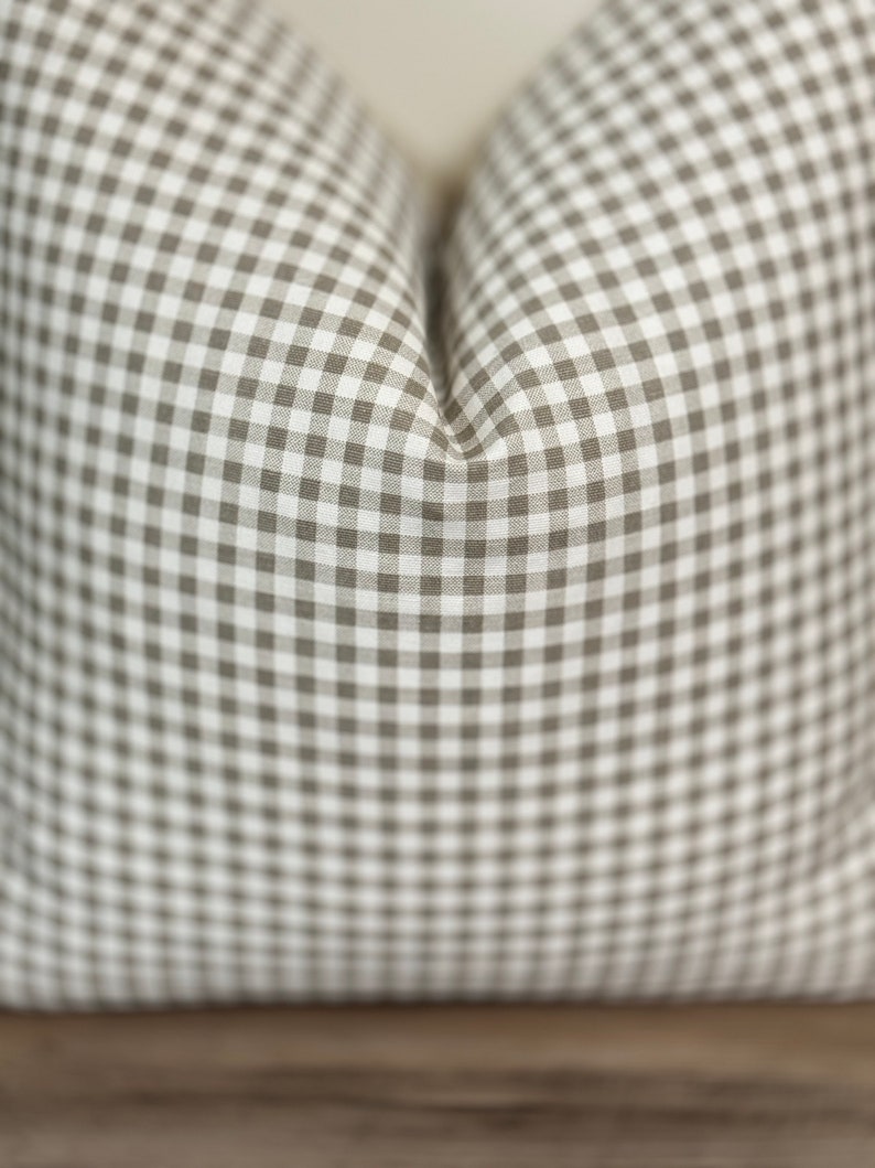 Puede incluir: Un coj&iacute;n cuadrado con un patr&oacute;n cl&aacute;sico de cuadros vichy en color topo y blanco. La tela presenta un dise&ntilde;o de cuadr&iacute;cula repetitivo a peque&ntilde;a escala con una apariencia suave y mullida, creando una hendidura marcada en el centro.