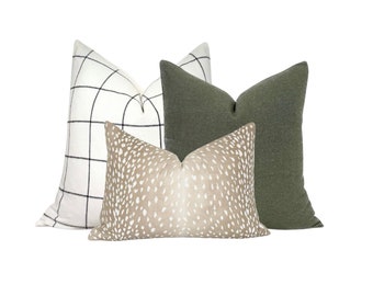 Puede incluir: Tres almohadas decorativas con diferentes patrones. La primera almohada es blanca con líneas de cuadrícula negras. La segunda almohada es de color marrón claro con un patrón de puntos blancos. La tercera almohada es de color verde oscuro.