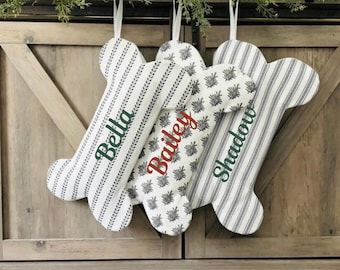 May include: Bone-shaped Christmas stockings with personalized names: Bella, Bailey, and Shadow. The stockings feature various patterns, including vertical stripes and a floral design, in white and gray. Each stocking has a white ribbon for hanging.