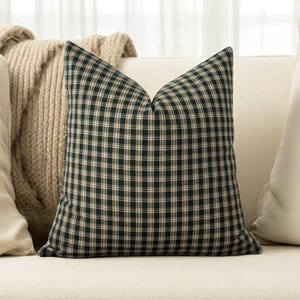 May include: A black and cream plaid throw pillow with a square shape. The pillow is sitting on a white couch.