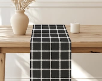 May include: Black and white checkered table runner. The runner is made of a woven fabric and has a subtle sheen. It is laid out on a wooden table.