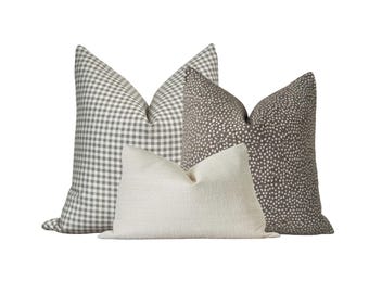 May include: Three decorative throw pillows. One pillow has a gray and white gingham pattern, another has a brown and white speckled design, and the third is a solid off-white color. The pillows are arranged together on a white background.